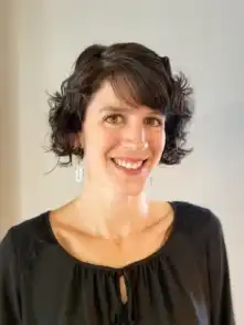 A smiling woman with short, curly dark hair wearing a black top and white earrings, posing against a plain background.