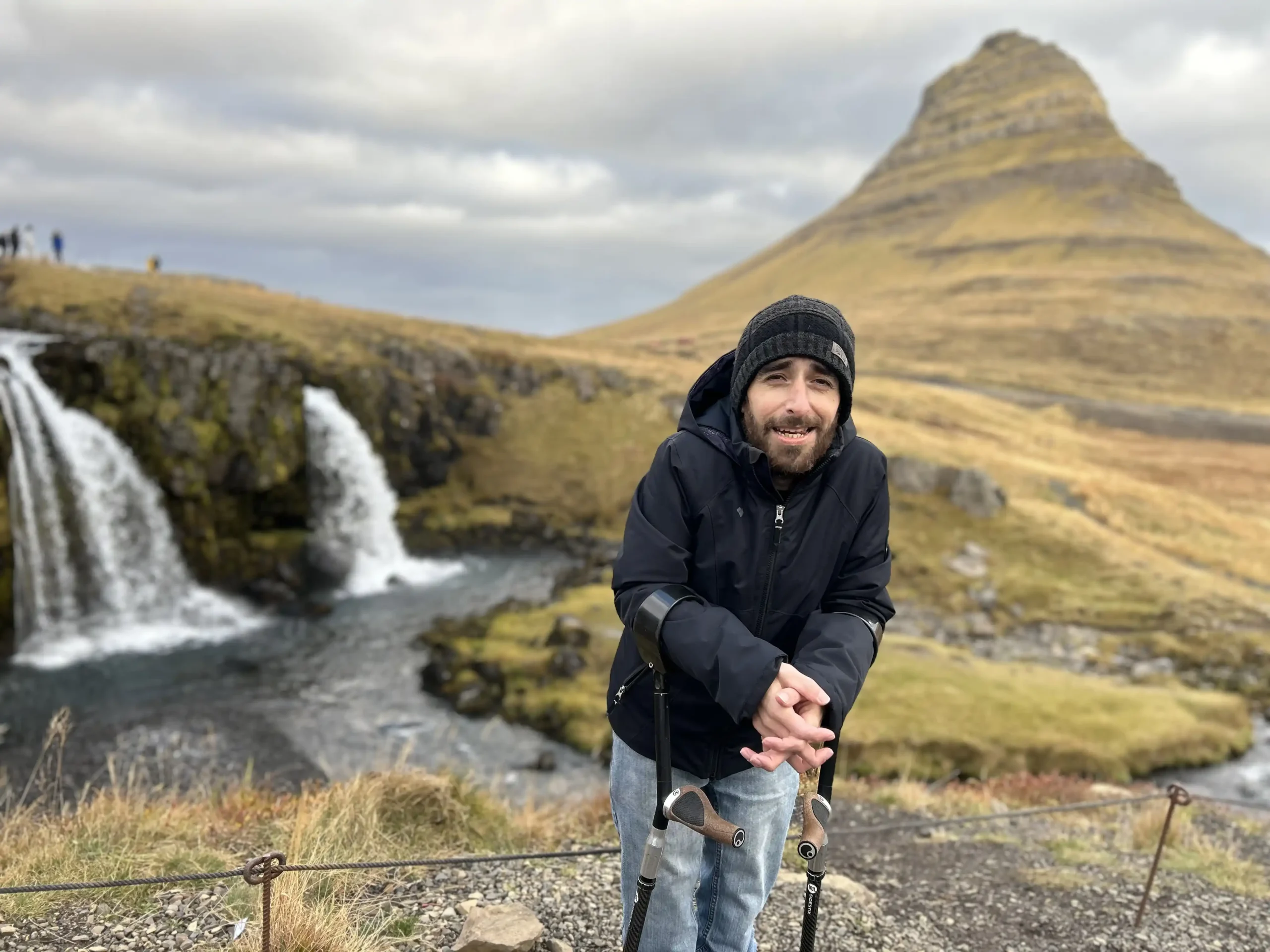 A man with diastrophic dysplasia, wearing a black jacket and a black beanie, is standing with the aid of crutches in front of a scenic landscape featuring a waterfall and a distinctive, steep, cone-shaped mountain in the background. The sky is cloudy, and the terrain around him is grassy with patches of rocky ground.