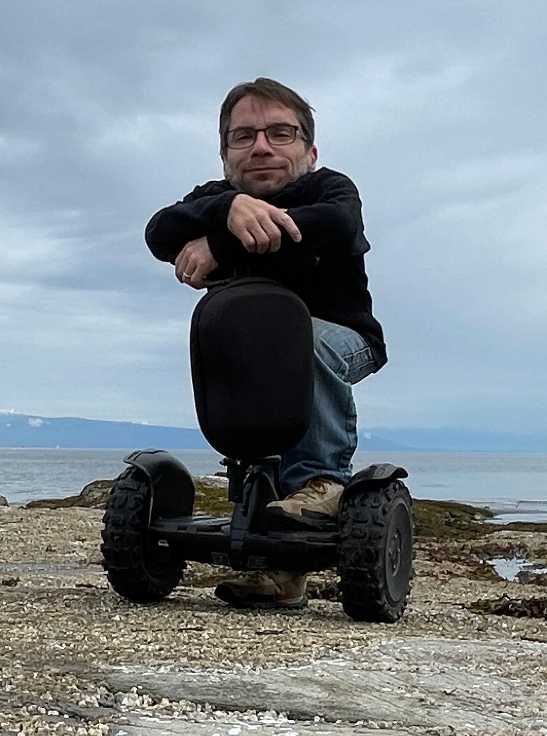 A man with glasses is seated on a Segway, resting his arms on the handlebars, and smiling at the camera. He is on a beach with a cloudy sky and mountains in the distant background. The terrain around him is rocky and covered with patches of sand. The atmosphere appears calm and serene.