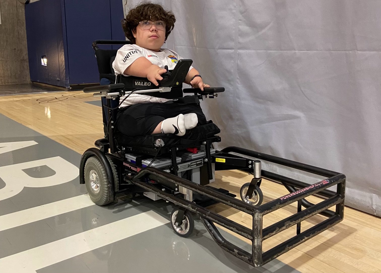 A young athlete sits in a specialized sports wheelchair on a gym floor. He has curly hair, wears glasses, and sports a white shirt with various sponsor logos. The wheelchair features a sturdy frame and protective bars at the front, designed for a specific type of sport. The athlete appears focused and ready for action, showcasing his determination and readiness to compete.