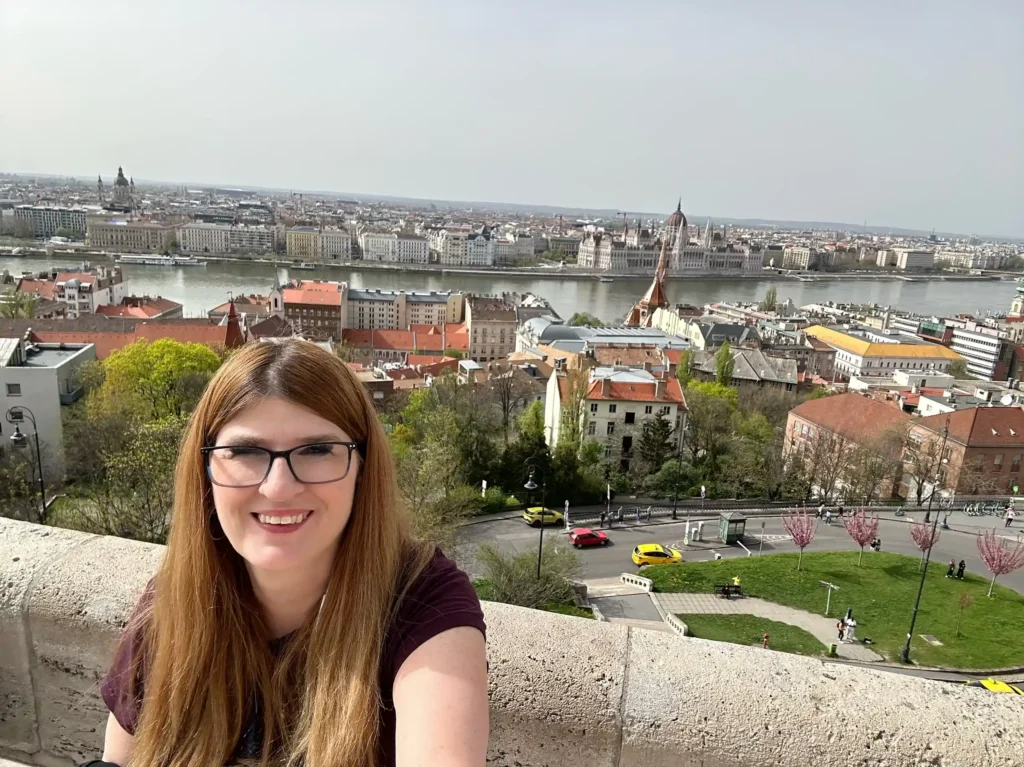 A woman with long, light brown hair and glasses is smiling while taking a selfie. She is standing on a high viewpoint overlooking a cityscape with a river running through it. The city has many buildings with red rooftops and historic architecture, including a prominent parliament building and a church. The scene is bright and clear, suggesting it is daytime with good weather. Trees and greenery are visible in the foreground, and the distant skyline is hazy but visible under a blue sky.