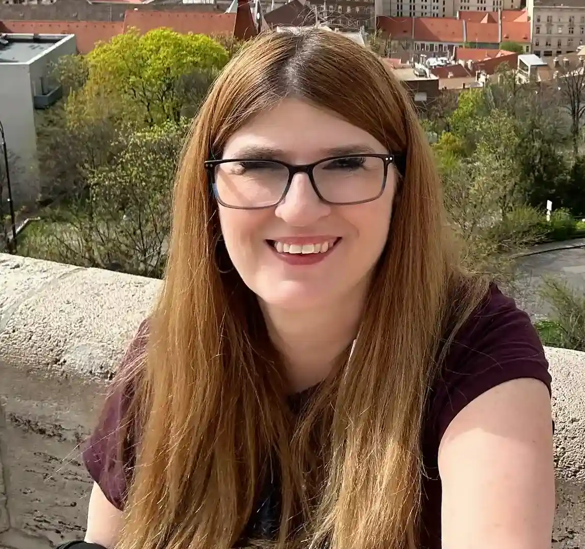 A woman with long, light brown hair and glasses is smiling at the camera while taking a selfie. She is wearing a dark shirt and is standing in front of a stone railing with a cityscape visible in the background. The background shows buildings with red rooftops, greenery, and part of a river. The overall scene is bright, suggesting it is a sunny day.