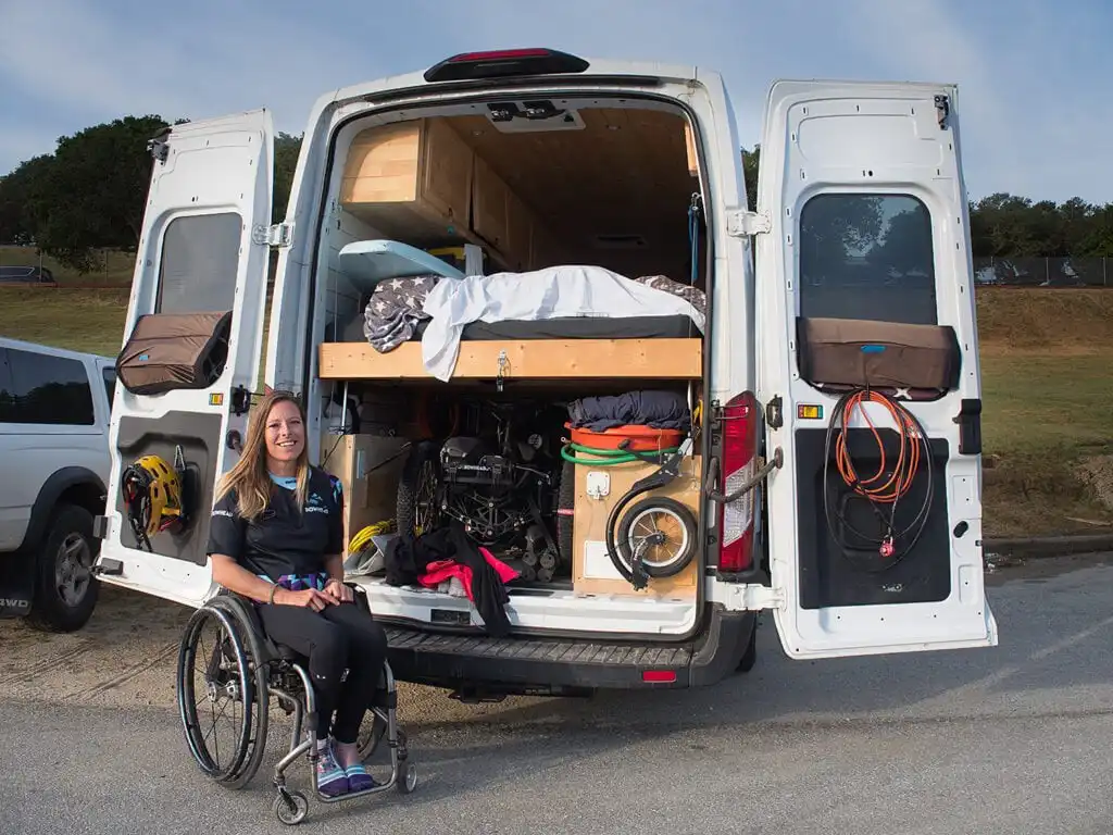A woman in a wheelchair is sitting next to the open back of a converted van. The van has its rear doors wide open, revealing a well-organized interior with a bed on a raised platform, various storage compartments, and equipment including a bicycle wheel and ropes. The woman is smiling and appears to be enjoying her mobile living setup. The setting seems to be an outdoor area, possibly a park or a camping spot, with a grassy field and trees in the background.