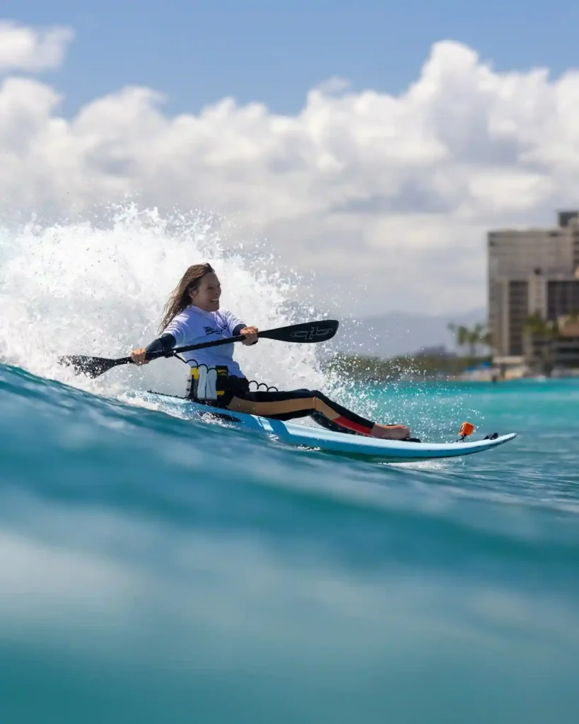 A woman with long hair is paddling on a modified surfboard in the ocean. She appears to be using a kayak paddle while riding a wave. The image captures her in action, with water splashing around her. She is wearing a long-sleeved white shirt and pants with a harness attached to the surfboard. In the background, there are buildings and palm trees, indicating a coastal location. The sky is clear with a few clouds, and the water is a vibrant blue.