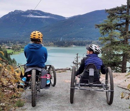 Two people are sitting in off-road wheelchairs, wearing helmets and outdoor clothing. They are on a trail overlooking a scenic lake with mountains in the background. The person on the left is wearing a blue jacket and a yellow helmet, while the person on the right is in a black jacket and white helmet. The scene suggests they are enjoying a peaceful outdoor adventure with a beautiful view.