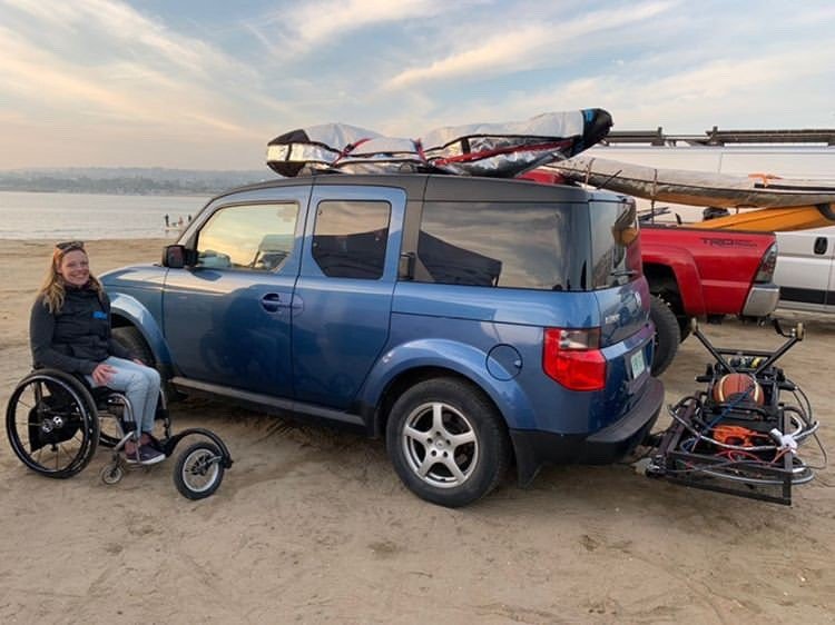 A woman in a wheelchair is sitting on the sand next to a blue SUV at the beach. The SUV has surfboards strapped to the roof and a rack attached to the back, carrying equipment. The beach and ocean are visible in the background, along with other vehicles and boats. The woman is smiling and dressed casually, enjoying the beach setting.