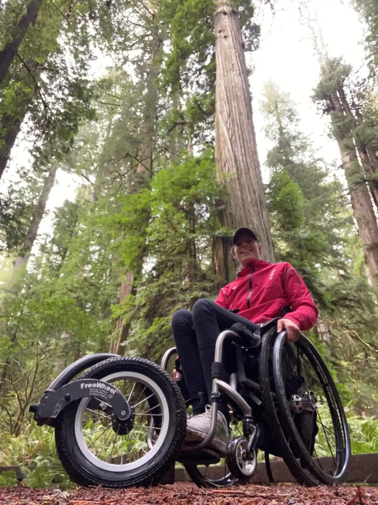 A person in a wheelchair is in a forest of tall, dense trees. They are wearing a red jacket and black pants, with a black cap on their head. The wheelchair is equipped with a large front wheel labeled "FreeWheel," which is designed for navigating rough terrain. The person appears to be enjoying an outdoor adventure, surrounded by the lush greenery and towering trees. The ground is covered in fallen leaves and twigs.