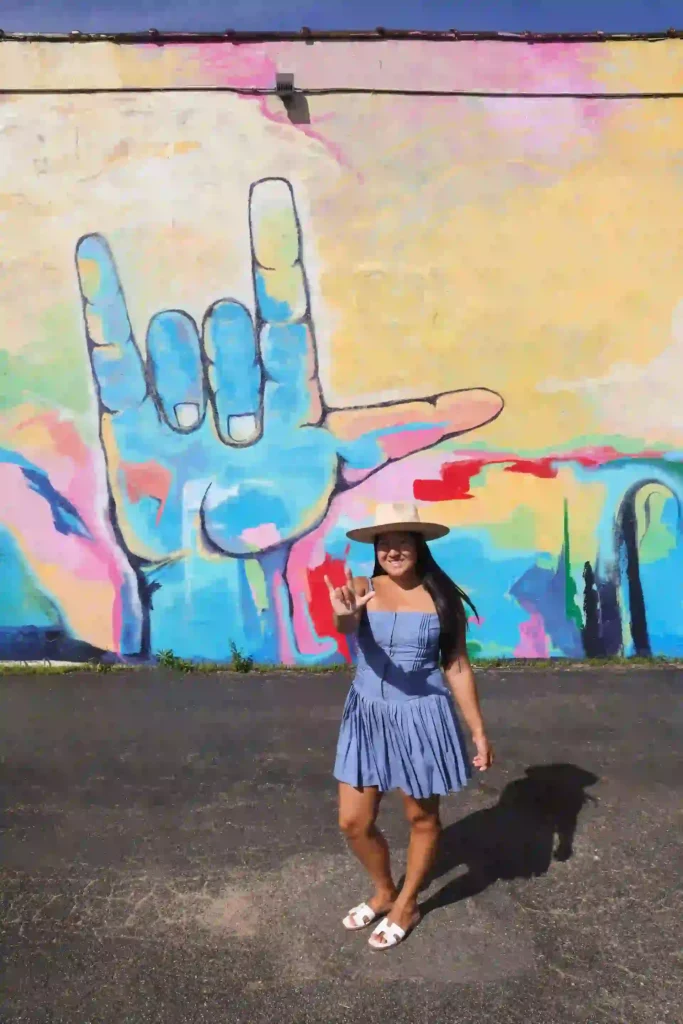 A woman is standing in front of a colorful mural. The mural features a large blue hand making the "I love you" gesture in American Sign Language. The woman is wearing a light blue dress, white sandals, and a wide-brimmed hat. She is smiling and also making the "I love you" gesture with her hand. The mural behind her is vibrant with abstract shapes and a variety of colors, including pink, yellow, red, and green. The ground is paved, and the sky above is clear and blue.