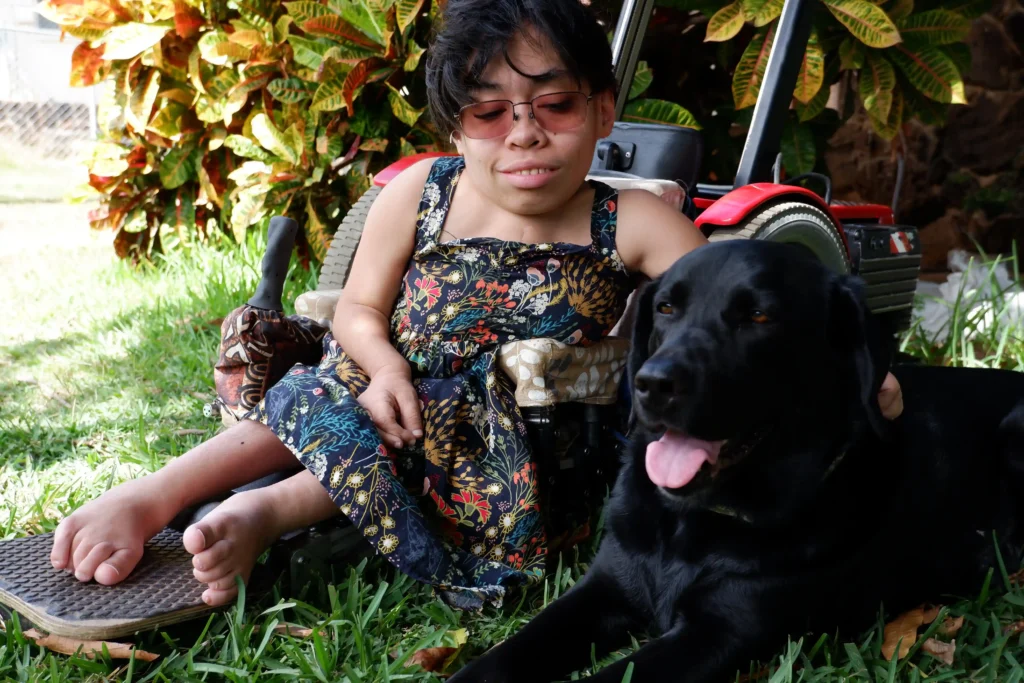 A woman in a wheelchair is sitting on grass with vibrant, leafy plants in the background. She is wearing a patterned dress with a floral and foliage design and sunglasses. Next to her is a black dog with its tongue out, looking content. The woman has short dark hair and is smiling gently while resting her hand on the wheelchair's armrest.
