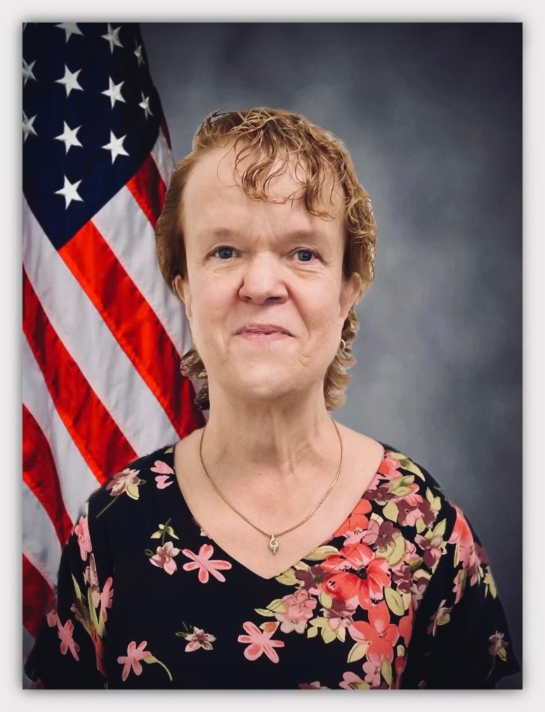 A professional portrait of a woman with short, curly hair, wearing a black floral blouse with pink and red flowers. She is standing in front of an American flag, which is partially visible on the left side of the image. The background is a gradient gray, giving the photo a formal and official appearance. The woman is smiling gently and wearing a small, elegant necklace.