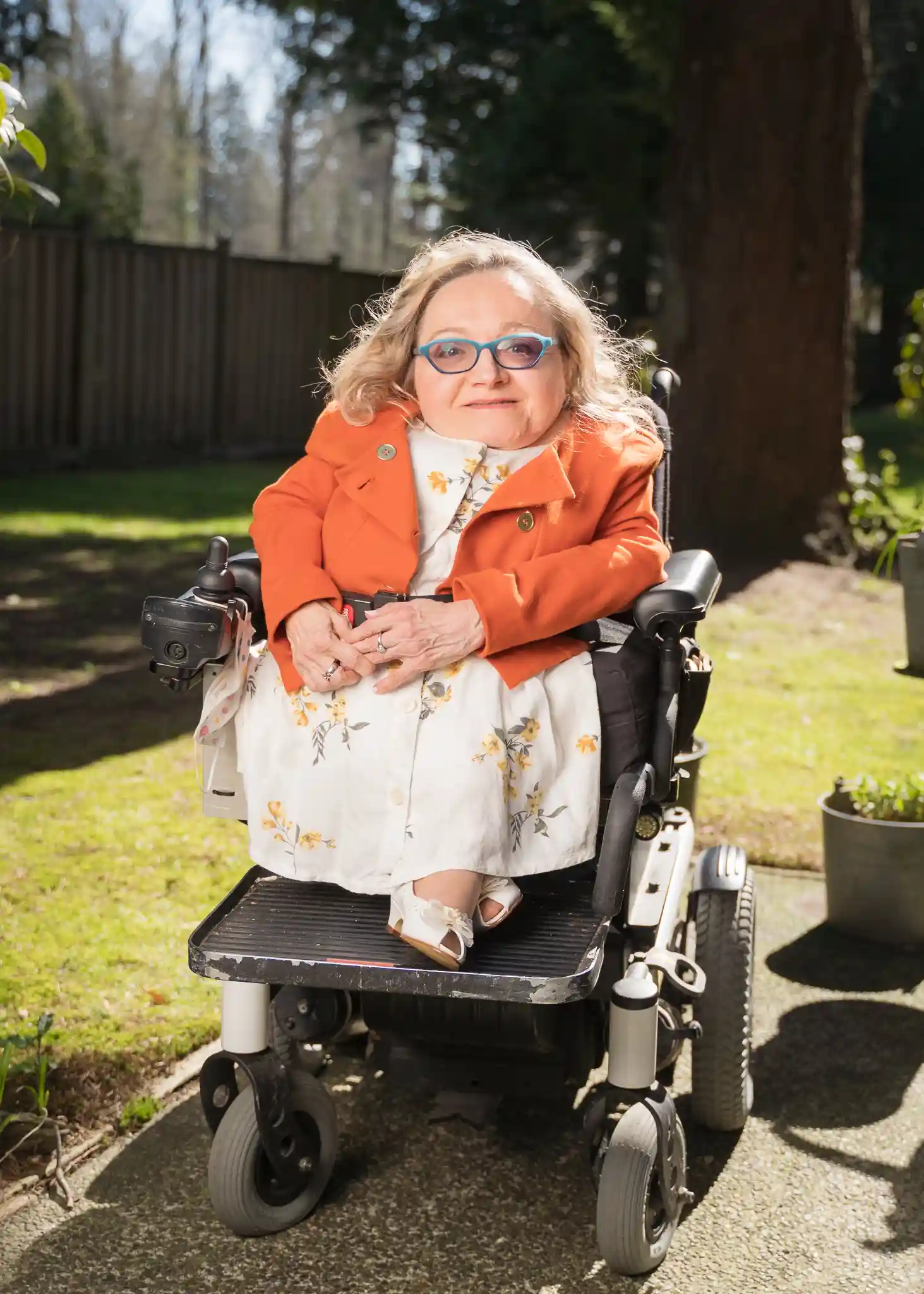 A woman in a motorized wheelchair is pictured outdoors on a sunny day. She has blonde hair and is wearing blue glasses, a white dress with a floral pattern, and an orange jacket. She is smiling at the camera, with a background of a wooden fence, green grass, and trees.