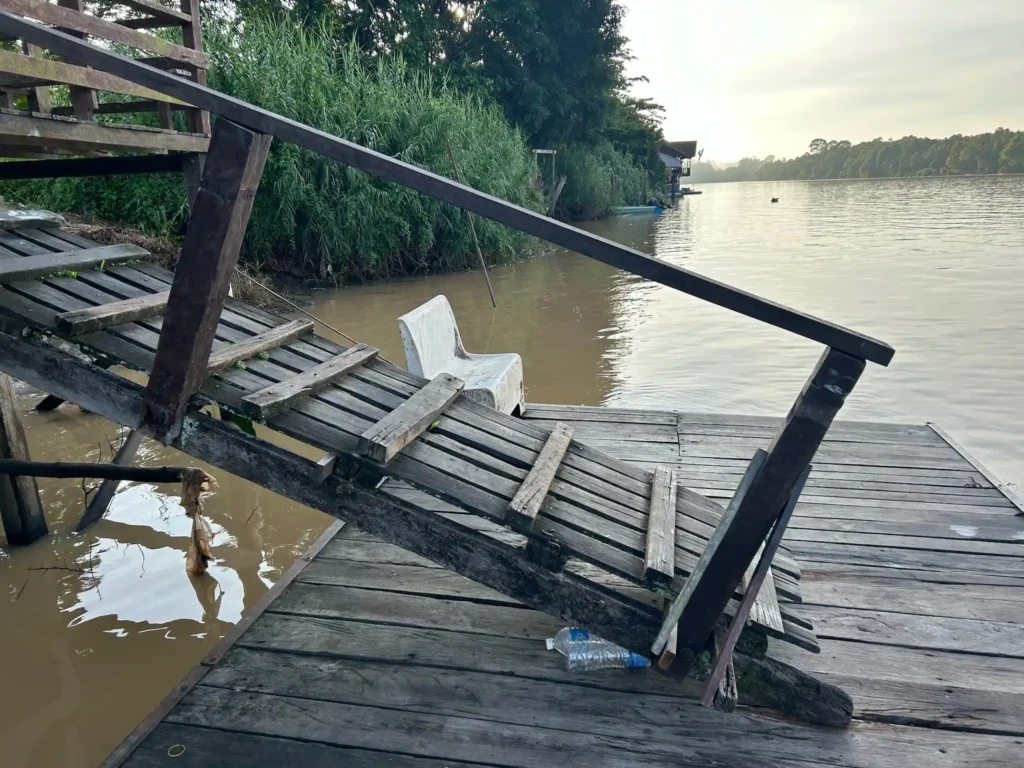 A wooden pier with a steep, old, and partially broken staircase leading down to a murky river. The pier has wooden planks, some of which appear to be damaged or worn out. In the background, there is dense green vegetation along the riverbank and a calm, wide river extending into the distance. There is also a plastic chair partially submerged in the water near the staircase. The overall scene suggests a rustic, possibly neglected riverside location.