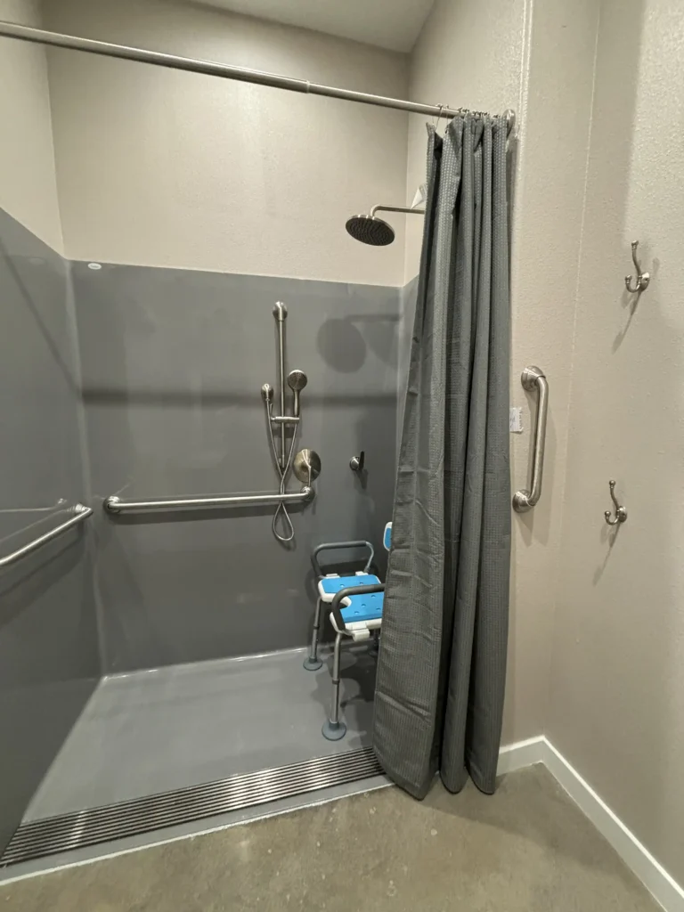 Roll-in shower: The image shows a roll-in shower designed for accessibility. It has a wide entry with no step, making it wheelchair accessible. Inside the shower, there are multiple grab bars mounted on the walls for support and stability. The showerhead is adjustable and mounted on a sliding bar, with an additional handheld showerhead attached to a flexible hose. There is a shower chair with a blue seat and backrest placed inside. A grey shower curtain is partially drawn, and there are additional grab bars and hooks mounted on the wall outside the shower area. The overall design is functional and accommodating for individuals with mobility challenges.