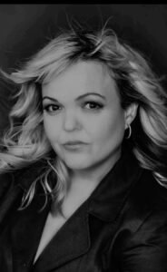 A black-and-white headshot of a woman with blonde, wavy hair. She has a serious expression, looking directly at the camera. She is wearing hoop earrings and a dark jacket. The photo has a high contrast with a sharp outline around her hair and upper body.
