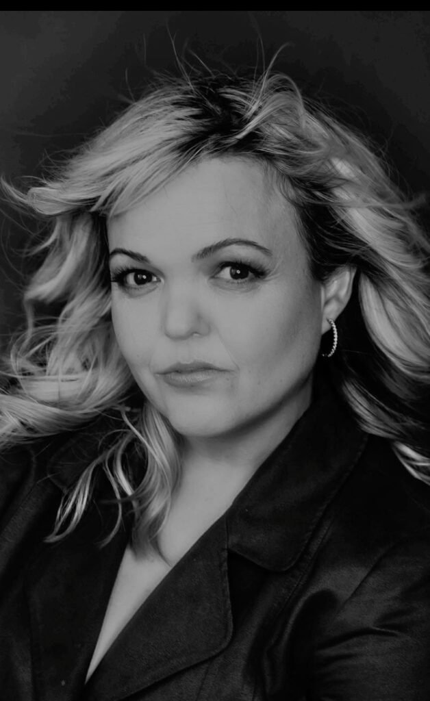 A black-and-white headshot of a woman with blonde, wavy hair. She has a serious expression, looking directly at the camera. She is wearing hoop earrings and a dark jacket. The photo has a high contrast with a sharp outline around her hair and upper body.