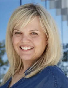 A smiling woman with shoulder-length blonde hair, wearing a blue top, standing in front of a blurred outdoor background