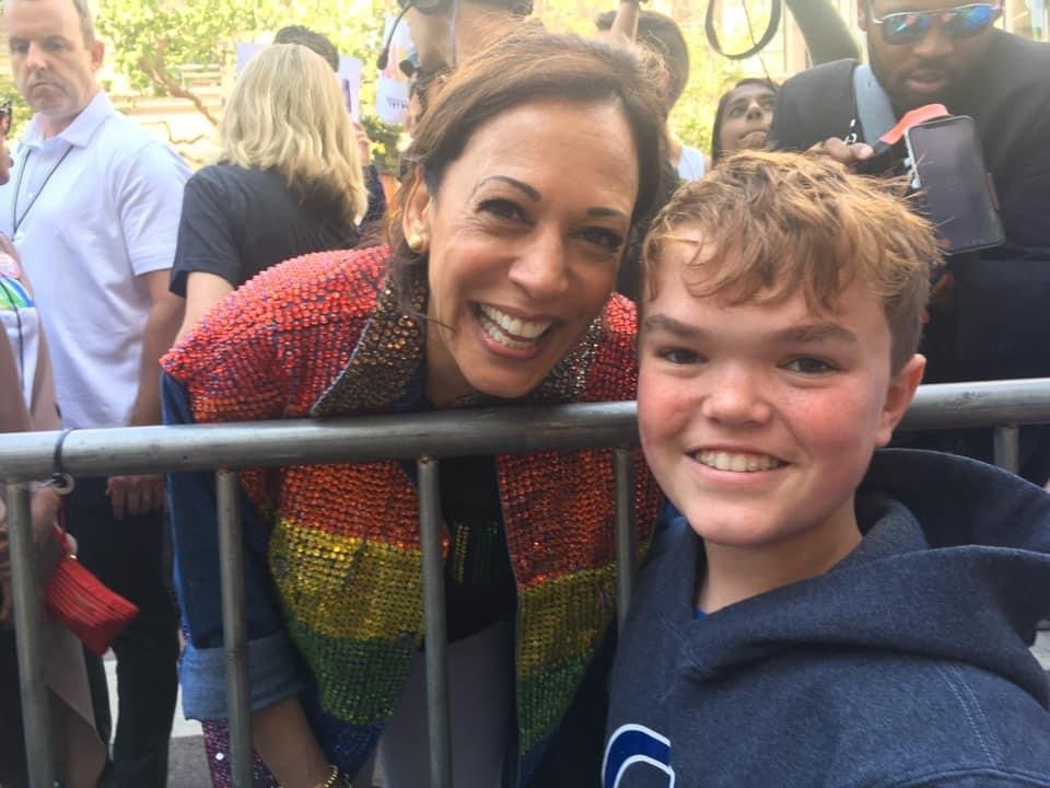 A woman and a young boy are smiling for a photo at an outdoor event. The woman, dressed in a colorful, sequined jacket with rainbow colors, is leaning over a metal barricade to pose for the picture. The boy is wearing a dark hoodie and standing in front of the barricade. Other people are visible in the background, some with cameras and phones. The atmosphere appears lively and festive.