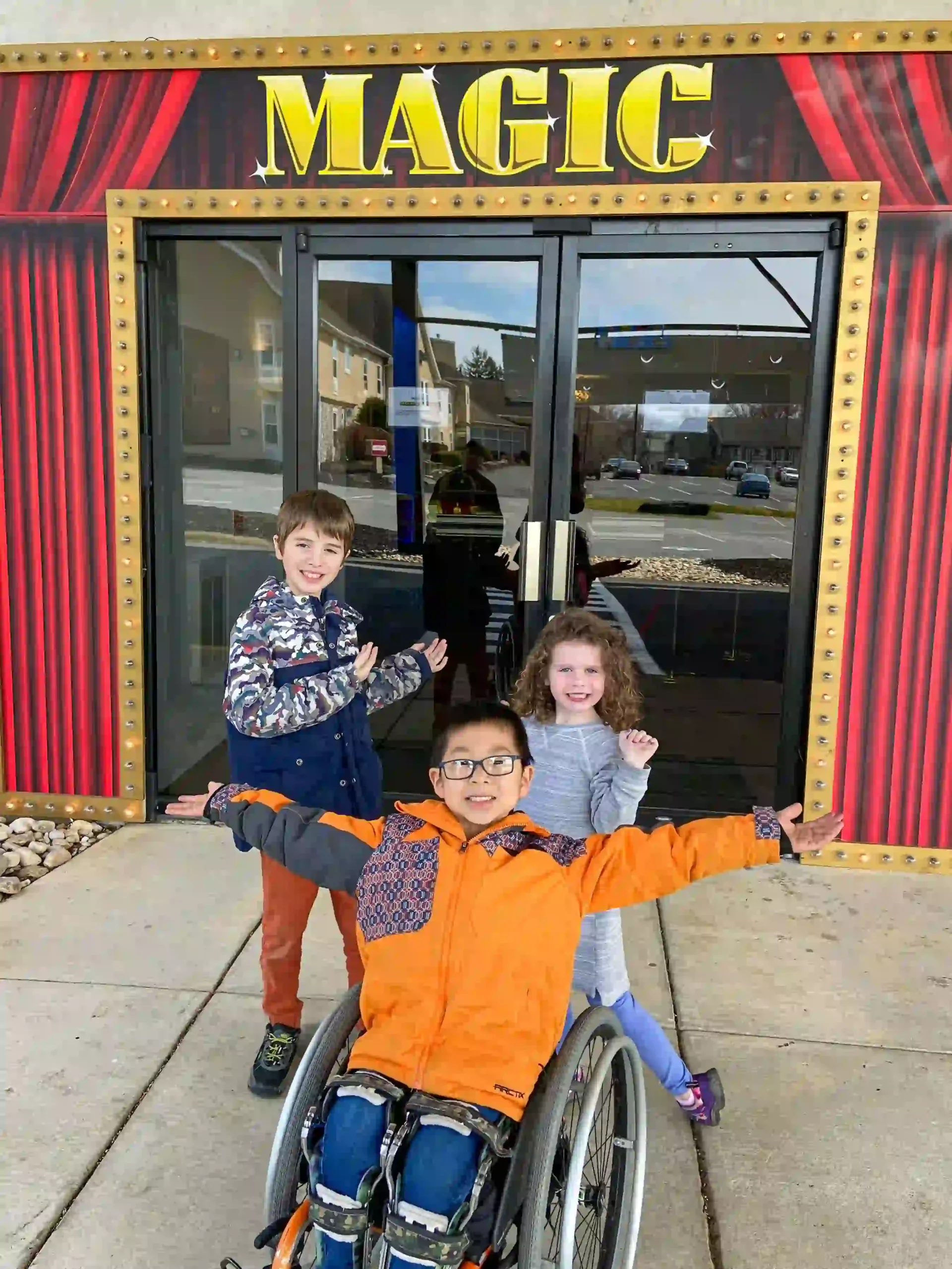 Three children are excitedly posing in front of a building with a large "MAGIC" sign. One child is sitting in a wheelchair with his arms outstretched, wearing glasses, a bright orange jacket, and jeans. To his left is a smiling boy in a blue and white patterned jacket with orange pants. To his right is a smiling girl with curly hair, wearing a gray dress and purple leggings. The entrance behind them has glass doors with reflections of the street and buildings visible. The building's facade is decorated with red curtains and gold trim, enhancing the magical theme.