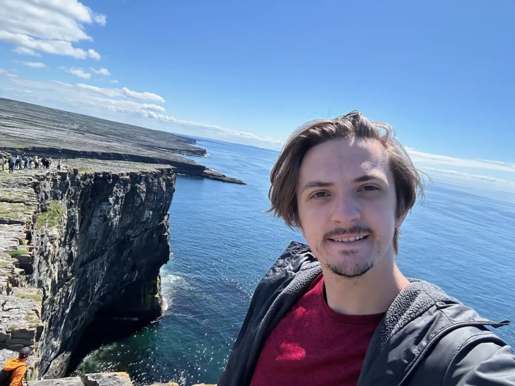 A young man with short brown hair and a slight mustache is taking a selfie on a sunny day near the edge of a cliff. The cliff overlooks a calm blue ocean, with other tourists visible in the background walking near the edge. The sky is clear with a few scattered clouds. The person is wearing a red shirt and a gray jacket. The location appears to be the Aran Islands, known for their rugged coastal scenery.