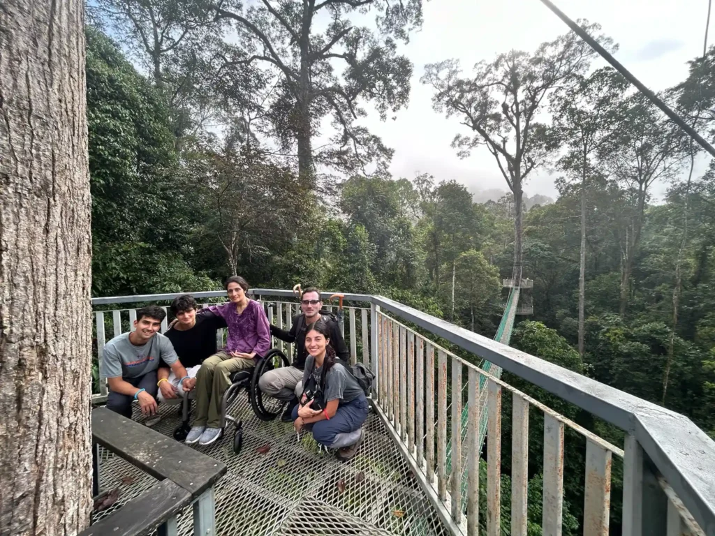 A group of five people is gathered on an elevated metal platform surrounded by lush, dense forest. The platform is bordered by metal railings, and a large tree trunk is visible on the left side. The group consists of four individuals sitting or kneeling on the ground and one person in a wheelchair. They are all smiling at the camera, and the forest canopy extends into the background, with tall trees and a misty atmosphere, indicating they are high up in the canopy of a tropical rainforest. A narrow suspension bridge extends from the platform into the forest.