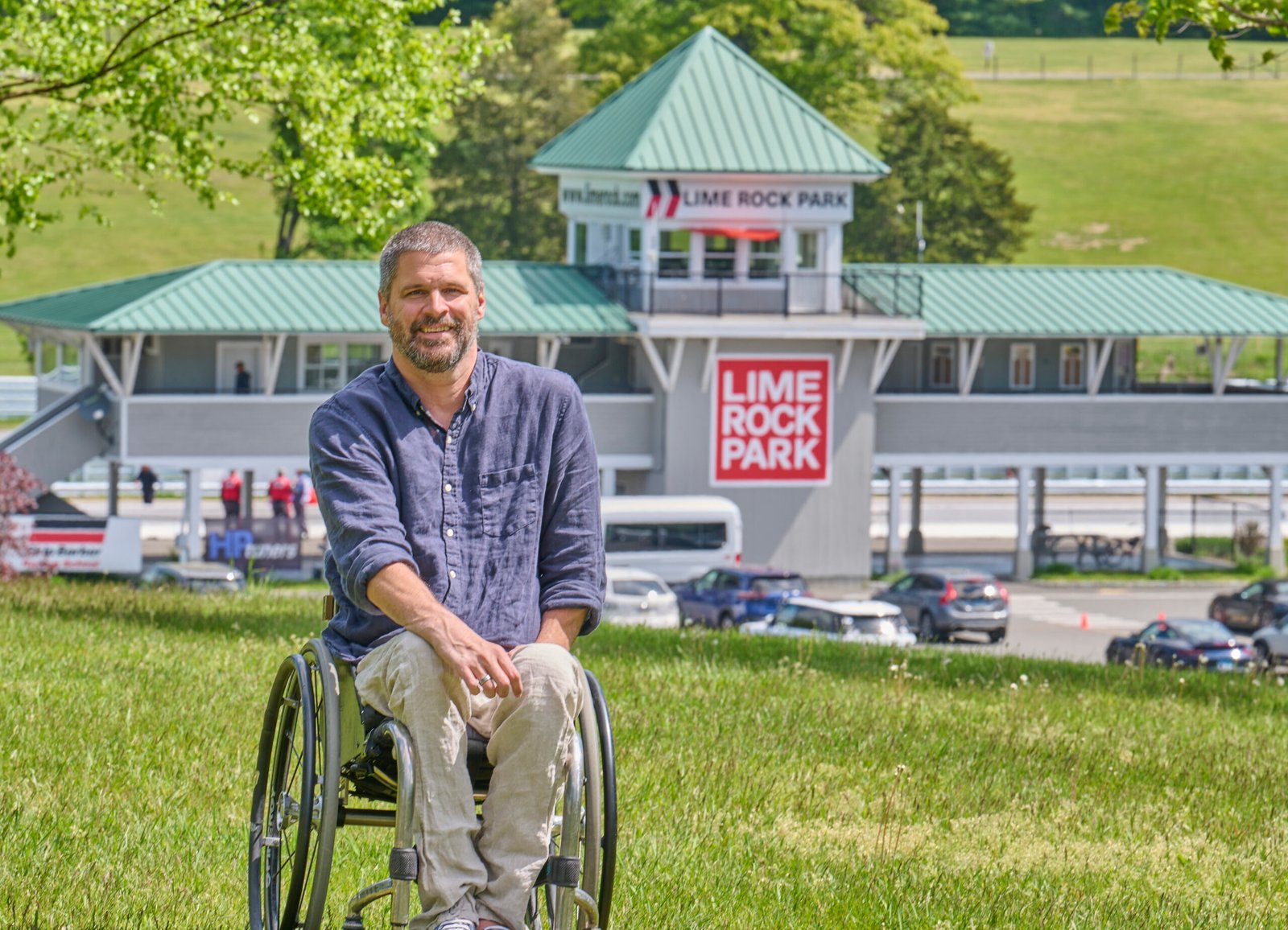 A man in a wheelchair is sitting on a grassy hill in front of the Lime Rock Park racetrack. He has short grayish hair and a beard, wearing a long-sleeve blue button-up shirt and beige pants. In the background, a two-story building with a green roof and a "Lime Rock Park" sign is visible, along with some parked cars and a few people walking around. The setting appears to be outdoors on a sunny day, with a relaxed and pleasant atmosphere.