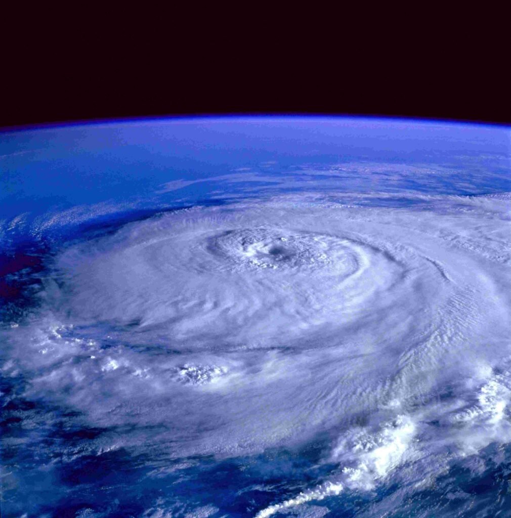The image shows a powerful hurricane captured from space, featuring a large swirling mass of white clouds forming a well-defined circular shape with a distinct eye at its center. The Earth’s deep blue atmosphere curves gently at the horizon, emphasizing the storm’s massive scale. The cloud formation spirals outward in intricate, turbulent bands, conveying the intensity of the natural phenomenon below.