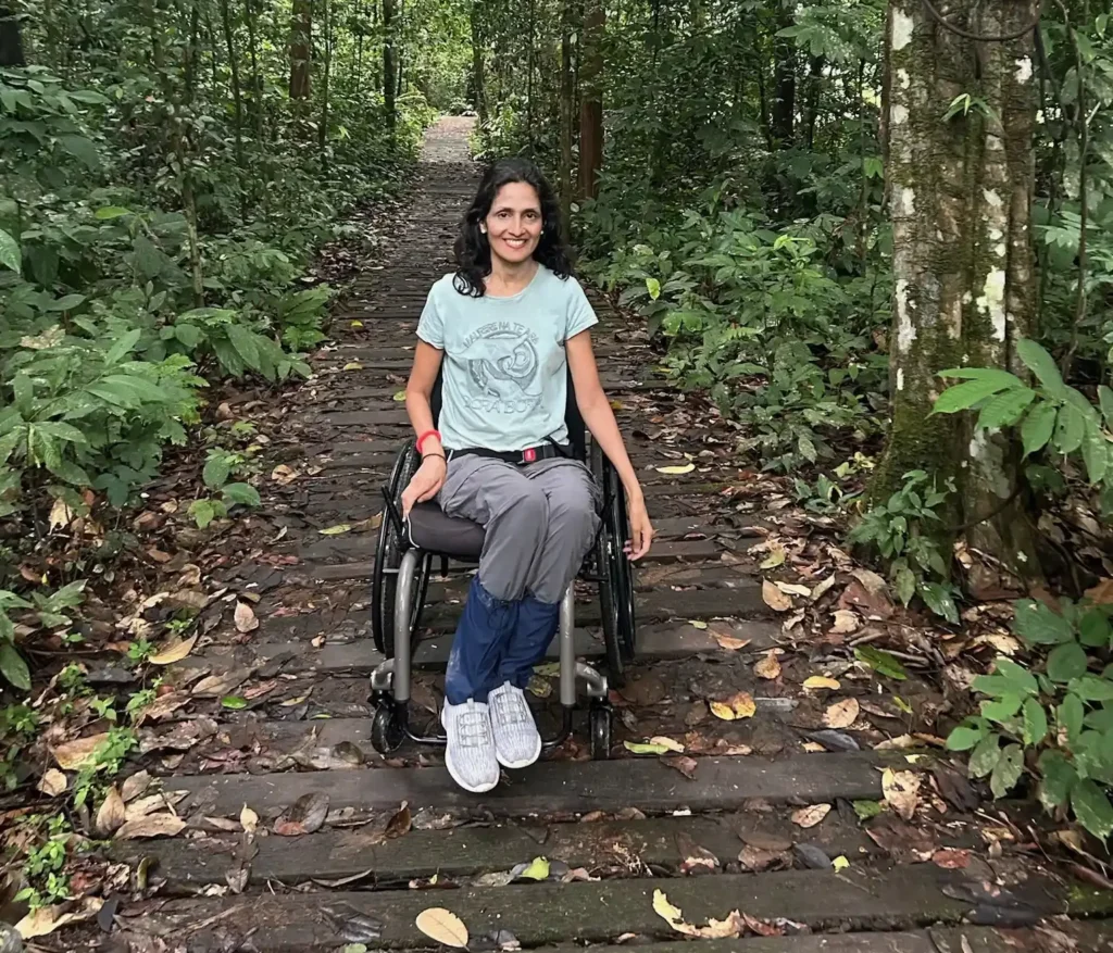 The image shows a woman with long, wavy dark hair and a bright smile. She is wearing a light-colored top, and the background appears to be an outdoor, nature setting with greenery and a path behind her.
