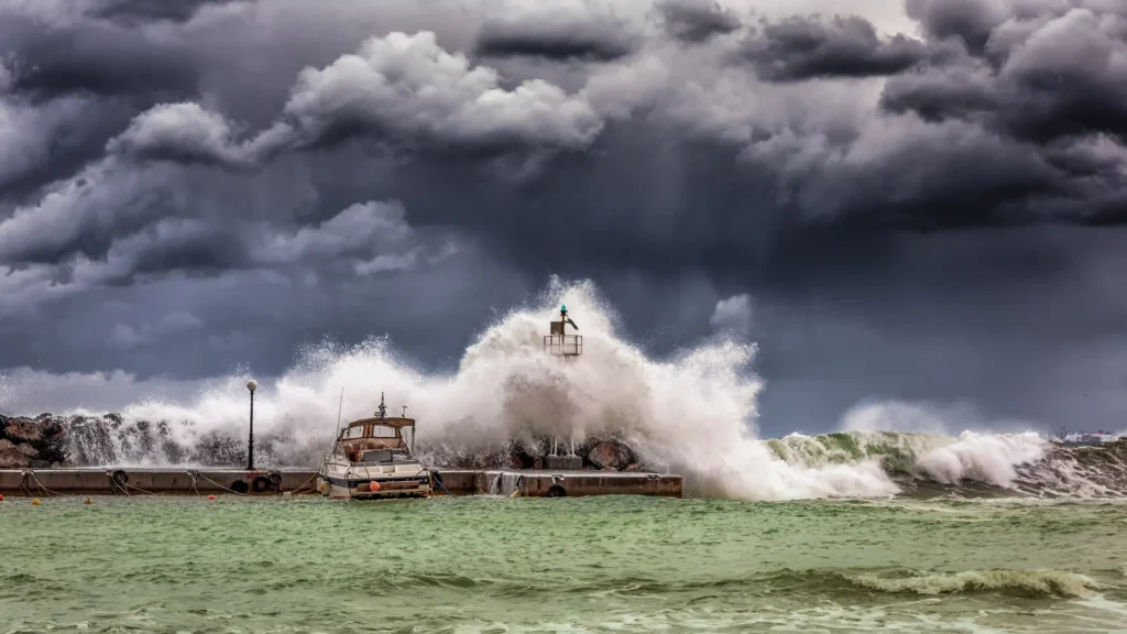 The image captures a dramatic scene of a storm with dark, ominous clouds looming overhead as massive waves crash against a dock. A small boat is moored to the side, seemingly vulnerable to the relentless force of nature. In the center of the dock, a lighthouse stands resolute as towering waves explode around it, spraying water high into the air. The sea churns in shades of green, reflecting the turbulence of the storm. The contrast between the stormy sky and the frothy waves creates an intense, powerful visual.