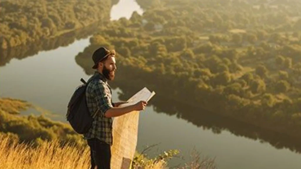 The image depicts a bearded man standing on a grassy hilltop overlooking a winding river below. He is dressed in a plaid shirt, a backpack, and a hat, holding a map in his hands as he gazes thoughtfully into the distance. The warm golden light of the setting sun bathes the scene, casting long shadows and illuminating the landscape, which is filled with dense trees and lush greenery. The peacefulness of the scene, combined with the sense of exploration, evokes a feeling of adventure and discovery in nature.