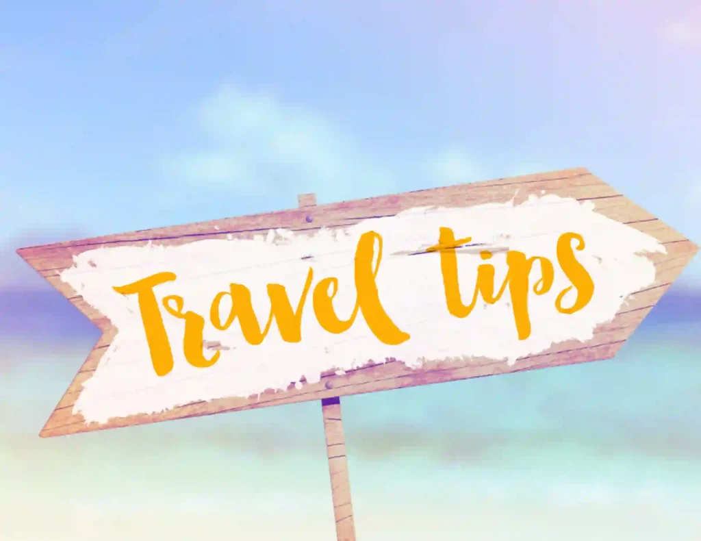 The image shows a wooden arrow sign with a worn-out, rustic texture. The sign has the words "Travel Tips" written in a bright yellow, cursive font. The background appears to depict a beach or tropical setting, with blue skies and blurred ocean waters in soft, pastel tones, creating a relaxed, vacation-like atmosphere.