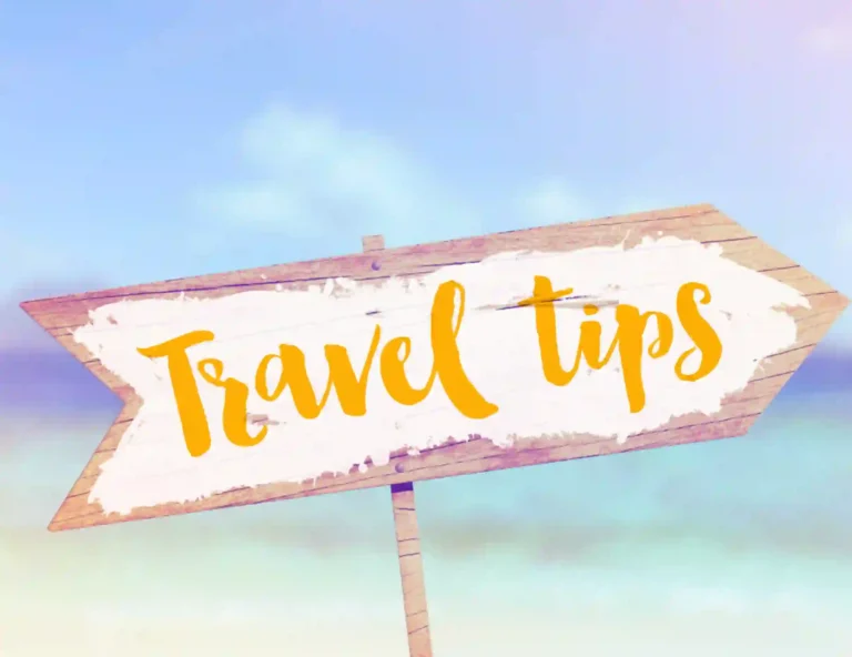 The image shows a wooden arrow sign with a worn-out, rustic texture. The sign has the words "Travel Tips" written in a bright yellow, cursive font. The background appears to depict a beach or tropical setting, with blue skies and blurred ocean waters in soft, pastel tones, creating a relaxed, vacation-like atmosphere.