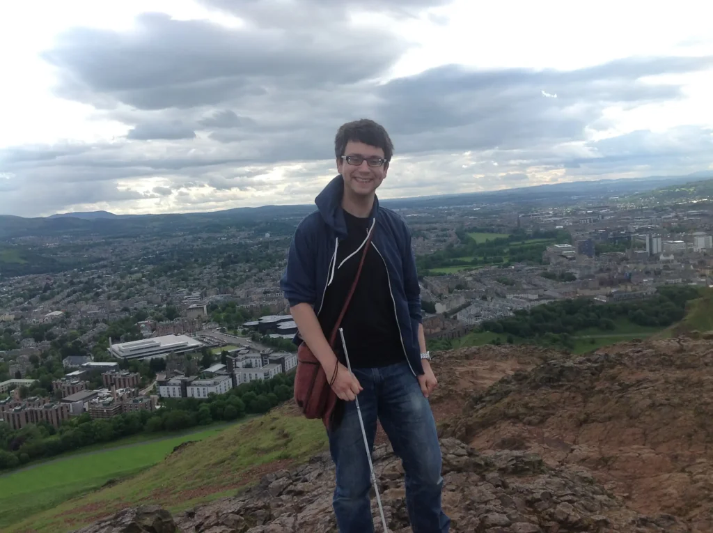 The image shows a person standing on a rocky hilltop, smiling at the camera. They are wearing glasses, a navy blue jacket, a black shirt, jeans, and carrying a crossbody bag. The person is also holding a white cane, suggesting they may be visually impaired. The background features a scenic view of a city below, surrounded by greenery and hills. The sky is cloudy with patches of sunlight breaking through. The location appears to be from a high vantage point, possibly Arthur's Seat in Edinburgh, UK