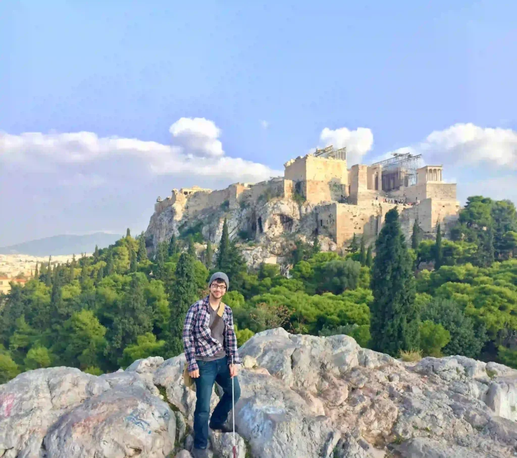 A person standing on a rocky hill with a backdrop of the Acropolis of Athens. The Acropolis, with its iconic ancient structures including the Parthenon, stands prominently on the hilltop. The surrounding area is lush with green trees under a bright blue sky, with some clouds. The person is wearing a checkered shirt, jeans, a gray beanie, and glasses, smiling as they pose near the rocks in the foreground.
