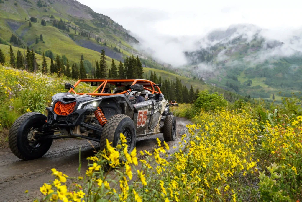 The image shows an off-road vehicle, possibly an all-terrain or utility task vehicle (UTV), driving through a scenic mountainous landscape. The vehicle has a sporty, rugged design with bright orange roll bars and shock absorbers, and it is marked with the number 55 on the side. It is driving on a dirt path surrounded by vibrant yellow wildflowers, with lush green hills and tall trees in the background. Misty clouds hover over the mountains, creating a picturesque and adventurous atmosphere. The scene appears to be part of a thrilling off-road rally or outdoor adventure.