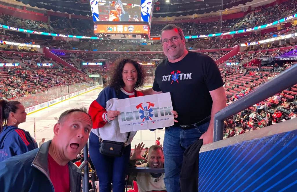 Three people stand together smiling at a hockey arena, with one person holding a '1st Tix' sign that reads 'Give Something to Those Who Gave.' The man on the right is wearing a '1st Tix' T-shirt, and the woman in the center is dressed in a red, white, and blue sweater. Another man photobombs with an open-mouthed expression from the front left. The background shows the ice rink and seated audience in the stadium.