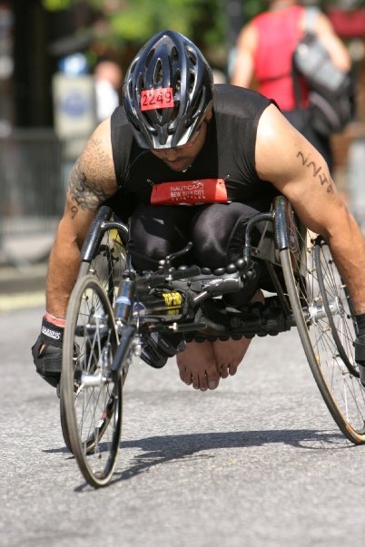 A determined athlete competes in the running portion of a triathlon using a racing wheelchair. The athlete, wearing a helmet and sleeveless black sports attire, propels himself forward with powerful arm strokes. His wheelchair is specially designed with aerodynamic wheels and a low frame for speed. A race number, 2249, is attached to the helmet, and spectators can be seen blurred in the background, highlighting the intensity of the race. The athlete's arms are tattooed, and his focus is visible as he moves down the course.