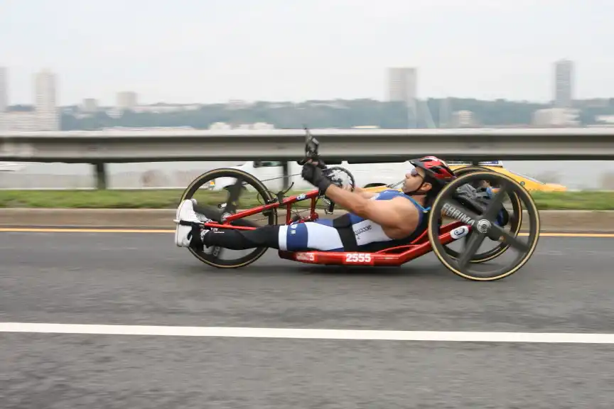 A handcyclist participating in a race, riding a red handcycle with a low reclining seat. The athlete is wearing a blue and white uniform, a helmet, and is focused on pedaling with their arms. The scene appears to be on a highway with a cityscape in the background, blurred by motion."