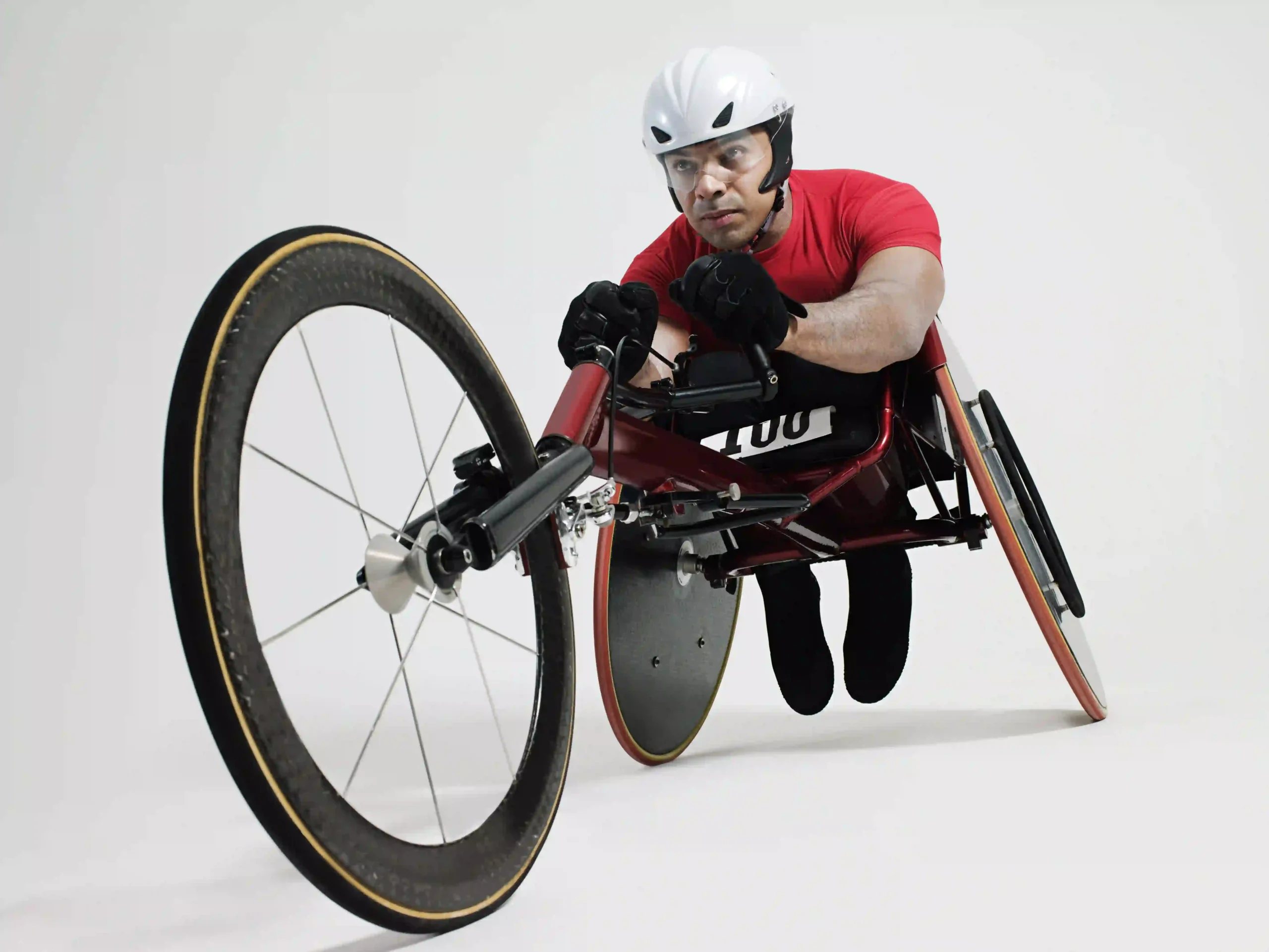 The image shows an athlete using a racing wheelchair, designed specifically for competitive events. The athlete is wearing a white helmet, a red shirt, and gloves, with a focused expression as they grip the handles of the racing wheelchair. The wheelchair has large, sleek wheels with thin tires, designed for speed and aerodynamic efficiency. The photo is taken from a low angle, emphasizing the front wheel and giving a dynamic perspective of the athlete in motion. The background is plain white, drawing full attention to the athlete and the equipment.