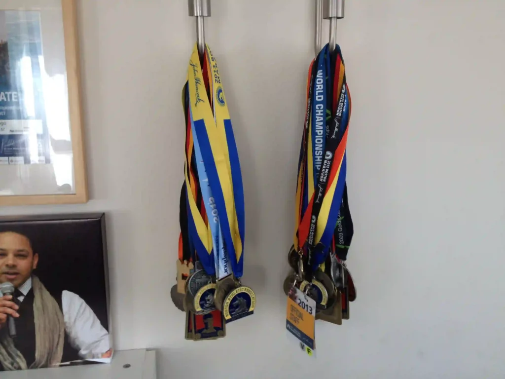 The image shows a display of two clusters of hanging medals, likely from athletic events such as races or championships. The medals are organized on metal hooks, with their colorful ribbons draped down. The left set includes a blue and yellow ribbon, while the right set features ribbons with the text "World Championship" and various other event names. Next to the medals is a framed image of a person holding a microphone, partially visible in the bottom left corner. There is also a visible badge from an event in 2013 hanging among the medals.