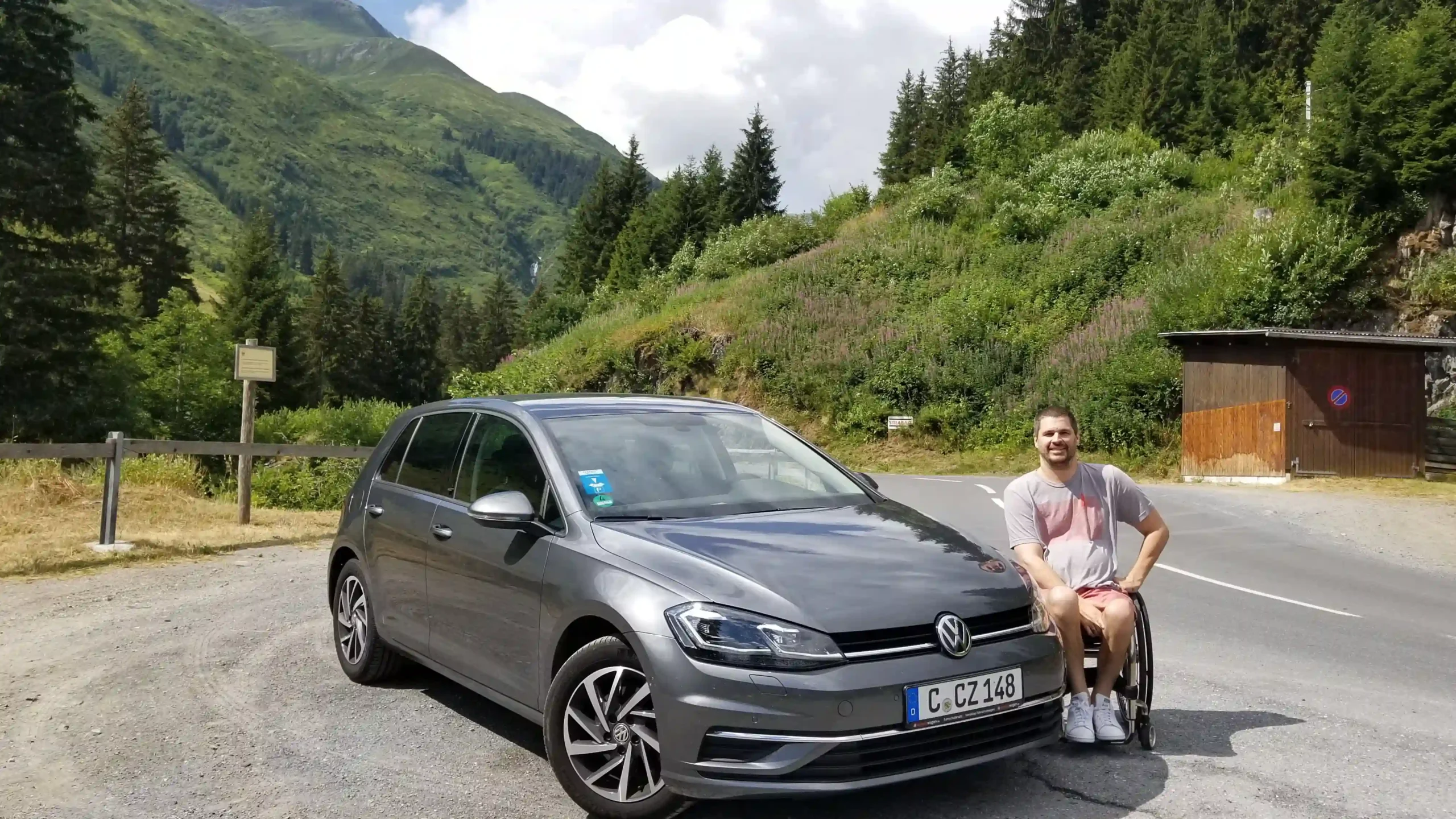 A man in a wheelchair is sitting next to a gray Volkswagen car with a German license plate on a mountain road. The background consists of lush green hills, dense forests, and a partly cloudy sky. The man is smiling, wearing a casual gray and red T-shirt, light shorts, and white sneakers. The setting appears peaceful, with a small wooden structure and signs visible along the road, suggesting a scenic stop in a mountainous area.