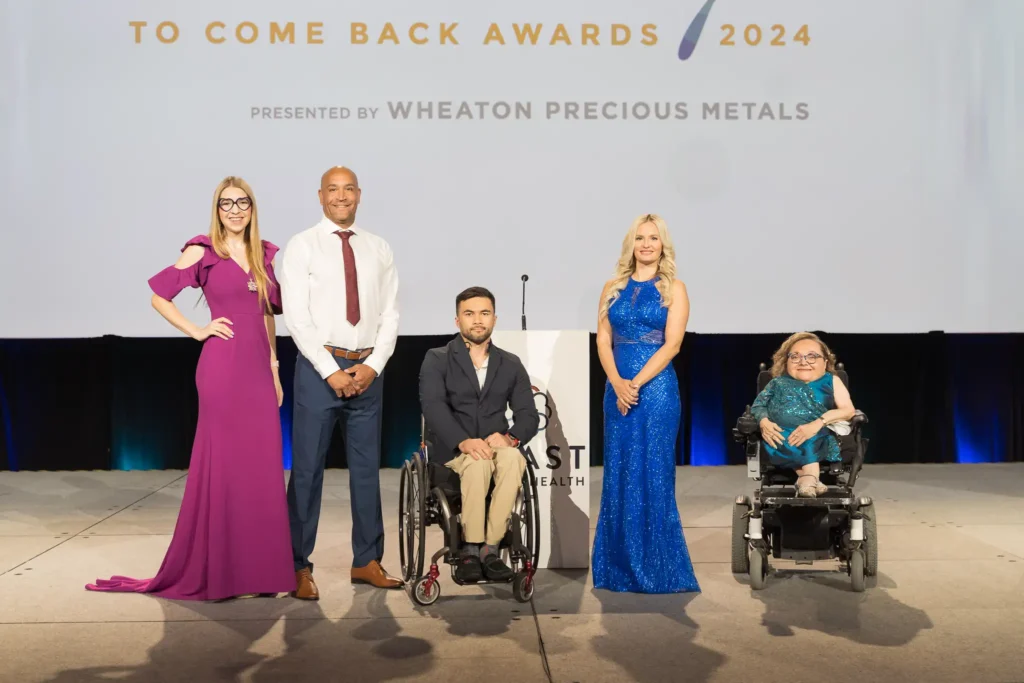 A group photo at the "Courage to Come Back Awards 2024" featuring five individuals standing on stage. From left to right: a woman in a purple gown with long straight hair and glasses, a man in a white shirt and tie standing confidently, a man seated in a wheelchair wearing a black suit jacket and beige pants, a woman in a sparkly blue gown with long blonde curls, and a woman in a motorized wheelchair wearing a teal-colored gown. The event backdrop shows the title "Courage to Come Back Awards 2024" with a sponsor logo "Wheaton Precious Metals."