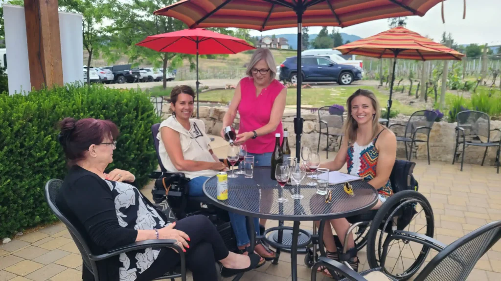 A group of four women is seated outdoors at a round table under large umbrellas, enjoying a wine tasting. Two of the women are in wheelchairs, smiling and engaging in conversation. A woman in a pink sleeveless top is standing, pouring wine into one of the glasses. The atmosphere is casual and relaxed, with a vineyard and parked cars visible in the background. The table has several wine glasses, a wine bottle, and a small can of soda. The setting seems to be a patio at a winery, with greenery and stone landscaping enhancing the ambiance.