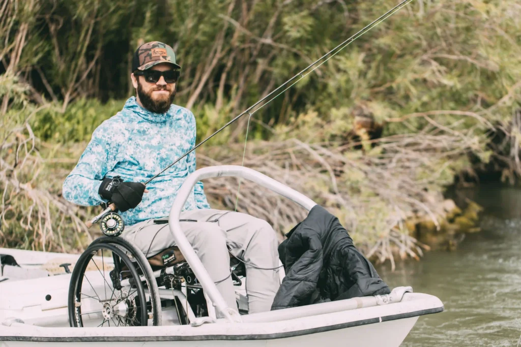 The image shows a man fishing from a boat. He is wearing a camouflage-patterned blue long-sleeve shirt, a camo hat, sunglasses, and gloves. The man is seated in a wheelchair, which is visible in the boat, and he is holding a fishing rod. Behind him, there is a backdrop of trees and foliage near the water. His posture and expression convey focus and enjoyment of the outdoor fishing activity.