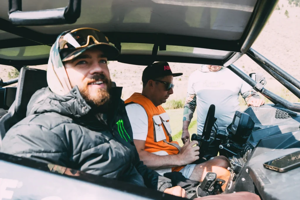 A group of men is seated inside an off-road vehicle. The man in the foreground is smiling, wearing a black jacket with a Monster Energy logo, a hood, a camo cap, and reflective sunglasses. Another man next to him is focused on the vehicle's controls, wearing an orange vest and a black cap with sunglasses. The third man, partially visible in the background, leans on the vehicle. The scene suggests they are preparing for or taking part in an outdoor adventure or motorsport activity, possibly in a rugged or mountainous terrain.