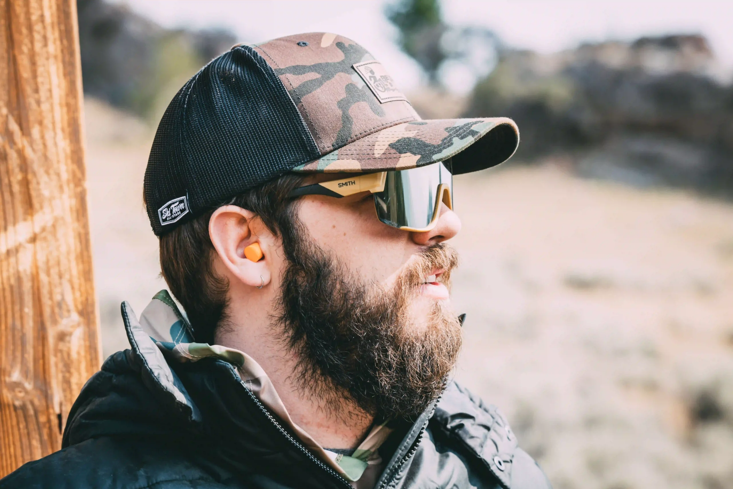 The image shows a bearded man wearing a camouflage-patterned cap, mirrored sunglasses with the brand "Smith" visible on the side, and orange earplugs. The man is outdoors, dressed in a black jacket with a high collar, and is looking off into the distance. The background is a natural, blurred outdoor landscape, giving the impression of a rugged or wilderness setting.