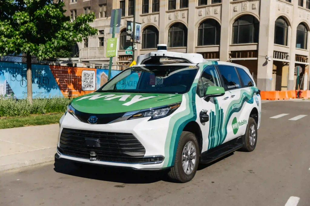 The image shows a green and white autonomous vehicle, branded with "MAV Mobility." The car appears to be a Toyota model, likely a minivan or SUV, with sensors and cameras mounted on the roof, indicating that it is equipped for self-driving technology. The vehicle is parked near a sidewalk, with a bus stop sign visible next to it. In the background, there are buildings with architectural details and construction signage. The design on the car features a cityscape and the words “ACCESSIBILITY,” emphasizing the mobility service's focus on accessibility.