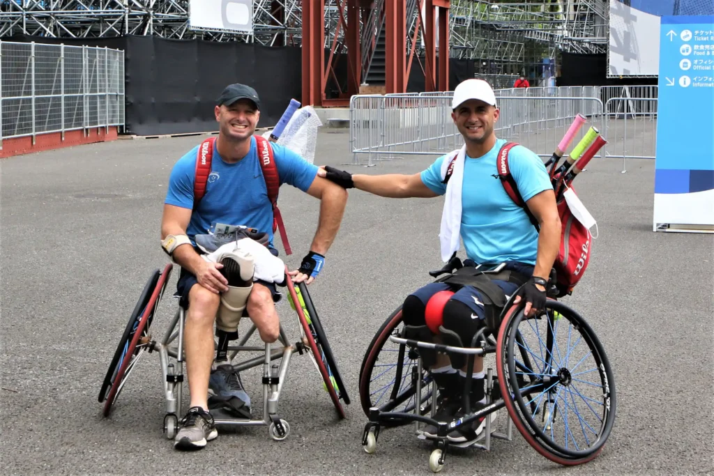 Two men in athletic gear, both seated in wheelchairs, are posing together outdoors on a paved area. They are both wearing blue shirts and have tennis racquets and sports gear attached to their wheelchairs. The man on the left, wearing a black cap, has a prosthetic leg visible and is smiling while resting one hand on his lap. The man on the right, wearing a white cap, extends his arm to rest a hand on his friend's arm. They appear to be at a sports venue, with metal barriers and a signboard visible in the background. Photo Credit: Credit -Keren Isaacson