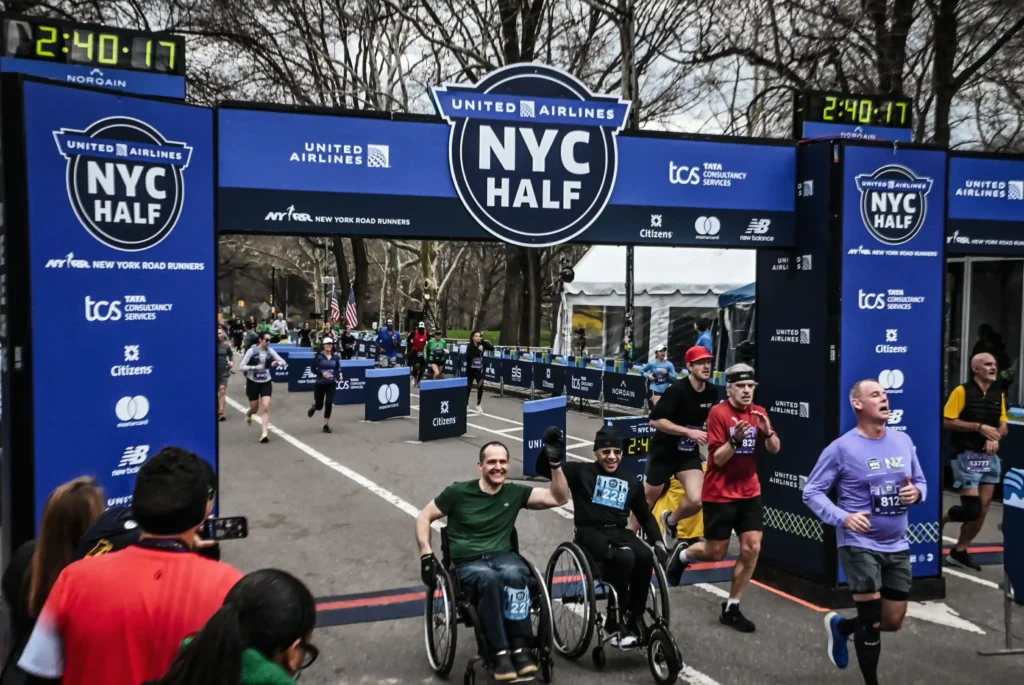 Image of the finish line at the United Airlines NYC Half Marathon. Two participants in wheelchairs are crossing the finish line, holding hands in celebration. A digital clock above the finish line displays a time of 2:40:17. Other runners are visible crossing the line, with some looking tired and others cheering. The event branding for the NYC Half Marathon is displayed prominently on the banner and the side panels, with sponsors like Tata Consultancy Services and New Balance also visible. Spectators are taking photos and cheering on the participants