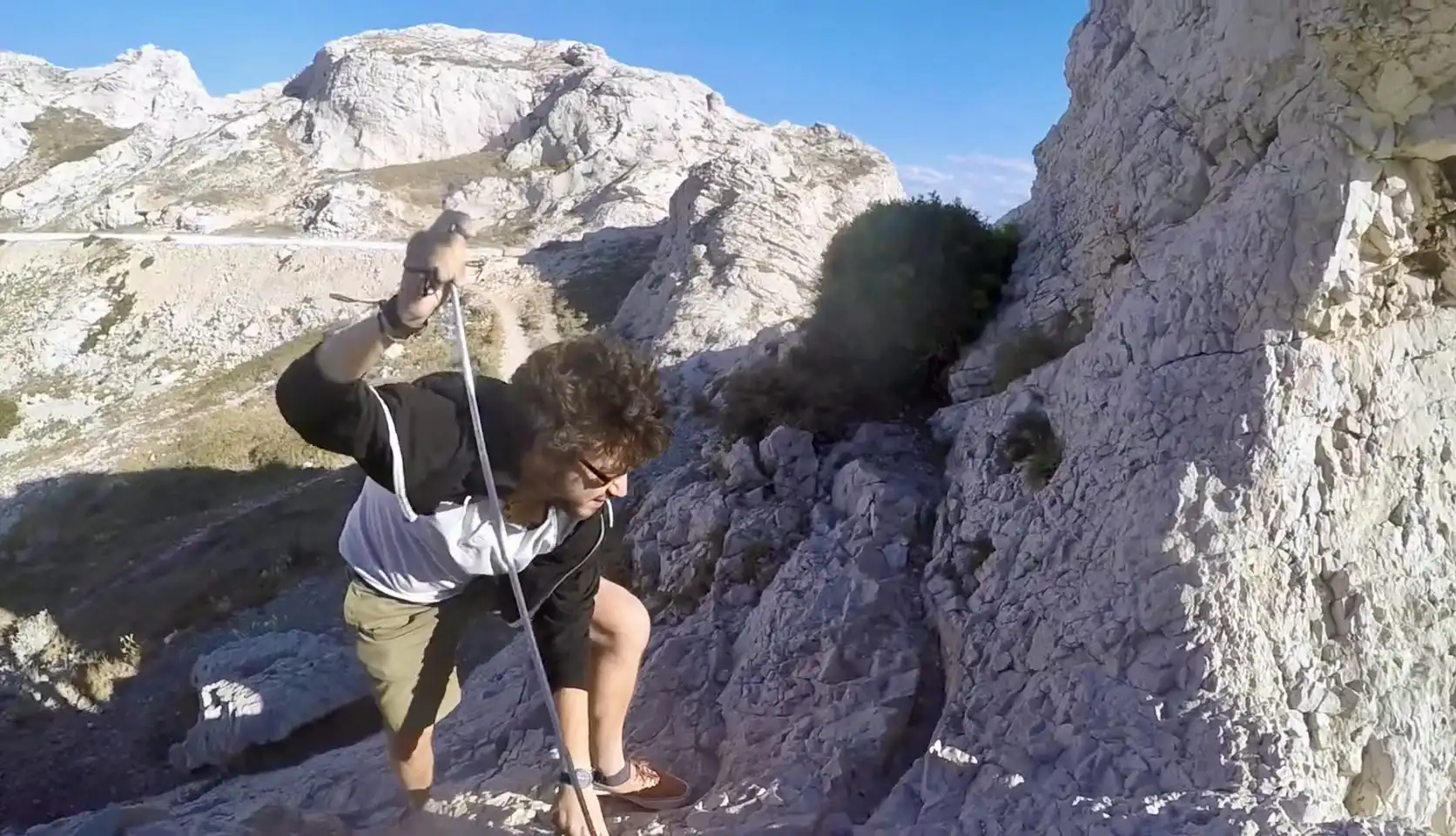 A person is climbing a rocky mountain face while holding onto a rope. They appear to be ascending a steep section of the rock, wearing a black and white long-sleeve shirt and shorts. The surrounding terrain is rugged, with white rocky cliffs and a mostly clear blue sky in the background. The climber is focused on their ascent, using the rope for support, and the landscape stretches into the distance, showing more mountainous formations and a valley below. The scene gives a sense of adventure and challenging outdoor activity.