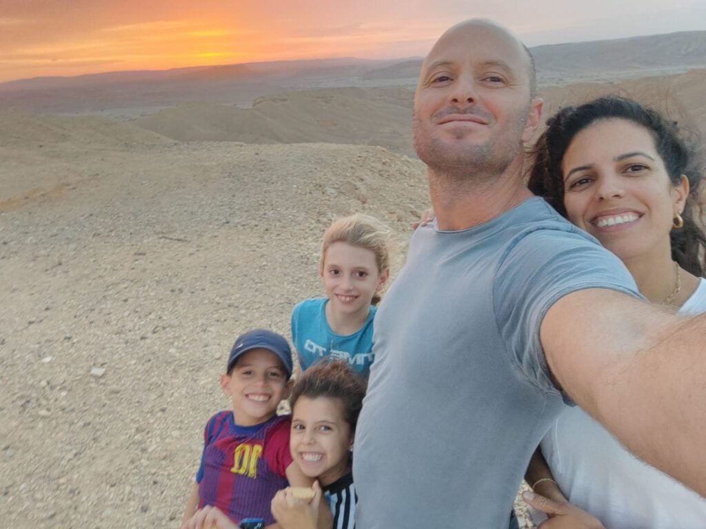 A family selfie taken outdoors at sunset with a rocky, desert landscape in the background. The group consists of two adults and three children. The man in the foreground is holding the camera, smiling with the woman next to him leaning close. The three children, standing in front, are smiling happily, with one wearing a cap and another wearing a blue shirt. The sky is a warm, golden hue with the sun setting behind the distant mountains, creating a serene and joyful atmosphere.